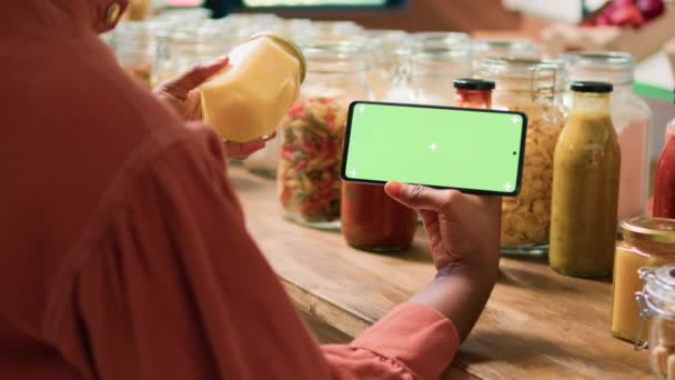 アフリカ系アメリカ人女性は 再利用可能な瓶に置かれた有機天然バルク製品をグリーンスクリーンを示しています 顧客は 単離された染色体ディスプレイでスマートフォンアプリをチェックします — ストック動画
