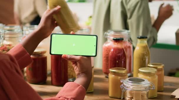 スマートフォンで緑色のディスプレイを持つ若い女性ショップは 地元の食料品店から大量に販売されている有機製品を選択します 地元で栽培されたエコフレンドリーな農産物を検索するビーガン顧客 — ストック動画