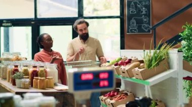 Çift organik biyolojik süpermarketteki ürünler hakkında soru soruyor, önlüklü depo sahibi taze ürün örnekleri sunuyor. Genç satıcı, sağlıklı beslenmeyi teşvik ediyor..