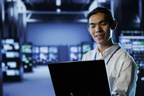 Smiling IT consultant in data center uses laptop to prevent system overload during peak traffic periods. Happy expert in server farm ensuring enough network bandwidth for smooth operations