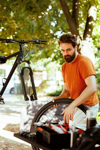 自宅の庭では 若い精力的な男性が損傷した自転車の車輪を交換するための専門的なツールを使用しています 快適なサイクリングのために外で分解された自転車のサービスは 献身的で健康的な白人男性のサイクリストです — ストック写真
