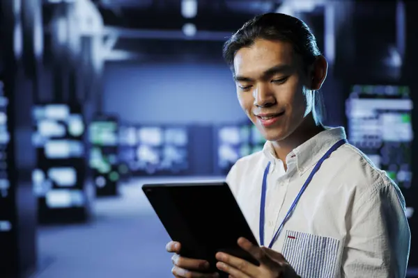 Cheerful computer operator in data center uses tablet to prevent system overload during peak traffic periods. BIPOC man in server facility ensuring enough network bandwidth for smooth operations