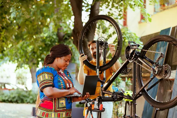 Sporty boyfriend and girlfriend repair bicycle damages in home yard using laptop. Active and vibrant man and black woman are exploring the internet for bike maintenance outside.