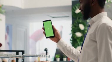 Premium giyim mağazasında şık kıyafetler satan Afrika kökenli Amerikalı bir adam cep telefonu tutuyor. Tasarımcı galerisinde yeşil ekranlı cep telefonu olan BIPOC müşterisi