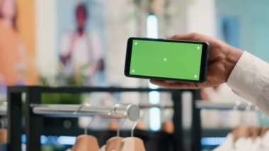 SH giyim mağazasındaki müşteri elinde yeşil ekran cep telefonuyla hangi kıyafetleri alacağına karar veriyor. Müşteri indirimli mağazada krom tuşlu akıllı telefon kullanarak yeni kıyafet koleksiyonuna bakıyor, kapat