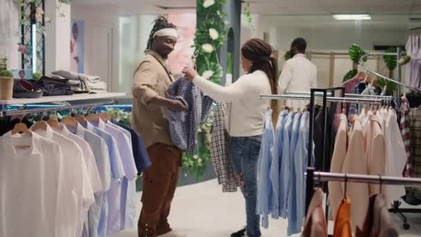 Bipocの男と彼のガールフレンドは 異なるシャツを試してセカンドハンドの衣料品店で安い服を探しています 低価格の衣服を検索する割引店で彼女のボーイフレンドと女性 — ストック動画