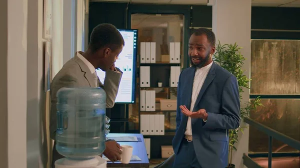 Happy african american coworkers enjoying nice water tank discussion during nightshift break. Smiling employee having cup of coffee relaxing while talking with colleague in office