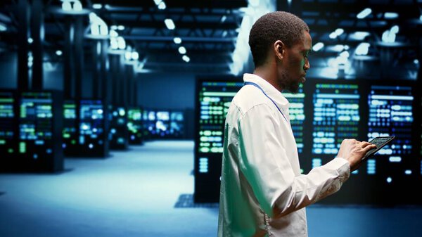 African american IT administrator investigating server clusters delivering web content to online customers. Precise serviceman using tablet to make sure critical systems are working properly