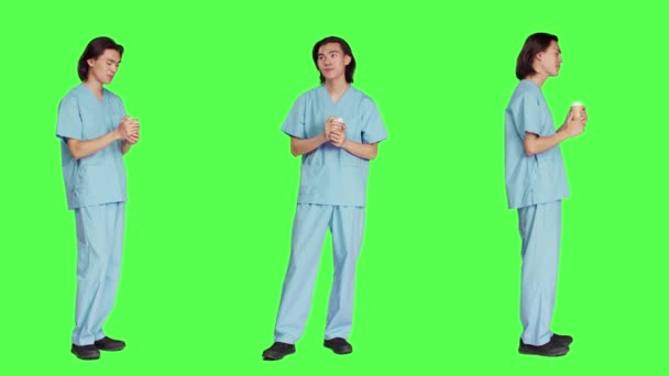 アジアのアシスタントは グリーンスクリーンバックドロップ カメラでホットカフェインリフレッシュを飲むヘルスケアスキルの男性スペシャリストにコーヒーを飲む ブルーユニフォームの医療看護師 — ストック動画