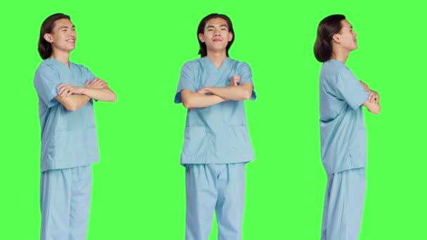 自信を持ってスタジオでポーズするフレンドリーな看護師 臨床服を着て 緑色の背景を越えた腕で立っています ヘルスケア業界で働く若手医療アシスタント — ストック動画