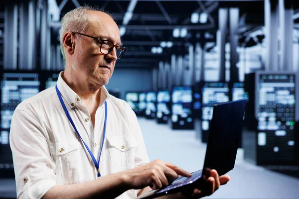 Senior IT developer inspects supercomputers in data center, ensuring smooth performance. Capable programmer in server farm monitoring energy consumption across units components using laptop