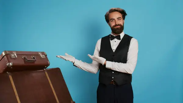 Hotel employee presents left side in studio, wearing formal suit and tie with white gloves while he does marketing ad, suggesting direction aside. Classy doorkeeper presents advertisement.