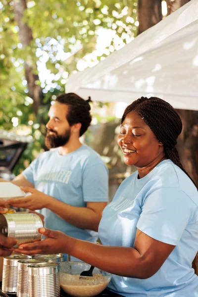 Close up image of african american female volunteer handing out canned goods to homeless people in need. Black woman and caucasian man share donation boxes filled with canned goods at food drive.