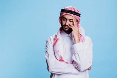 Thombe ve Gutra başörtüsü takmış düşüncelere dalıp tapınağı tutan düşünceli Müslüman adam. Geleneksel kıyafetler giyen bir Arap stüdyoda düşünceli bir yüz ifadesiyle poz veriyor.