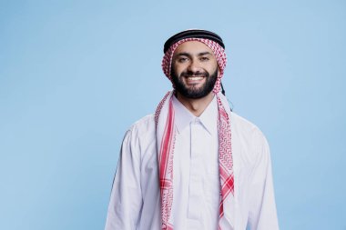 Geleneksel Müslüman kıyafetleri giymiş gülümseyen bir adam neşeli bir stüdyo portresiyle ayakta duruyor. Kameraya kaygısız duygularla bakarken başörtüsü ve başörtüsüyle poz veren mutlu bir Arap.