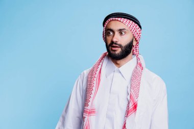 Geleneksel Müslüman kıyafetleri giyen adam şok olmuş yüz ifadesini görmezden geliyor. Arap bir adam tobe ve başörtüsü takmış, şaşırmış bir şekilde stüdyoda poz verirken ağzı açık duruyor.