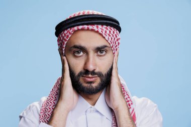 Müslüman adam kulaklarını palmiyelerle kapatırken kötü bir şey duymuyor. Üç bilge maymun stüdyo portresi konsepti. Geleneksel giysiler giyen bir Arap, işitme sorunu yaşıyor ve kameraya bakıyor.
