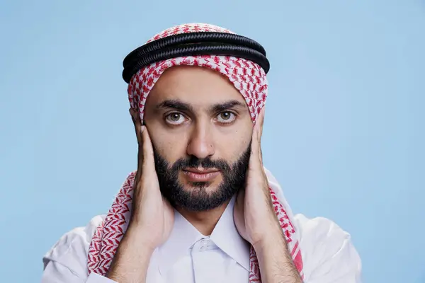 Muslim man closing ears with palms while showing hear no evil three wise monkeys concept studio portrait. Arab person wearing traditional clothes showcasing hearing problems and looking at camera