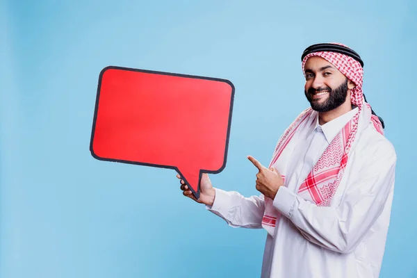 Smiling muslim man holding red speech bubble mockup and pointing with finger portrait. Person dressed in arab traditional clothes standing with message banner and looking at camera