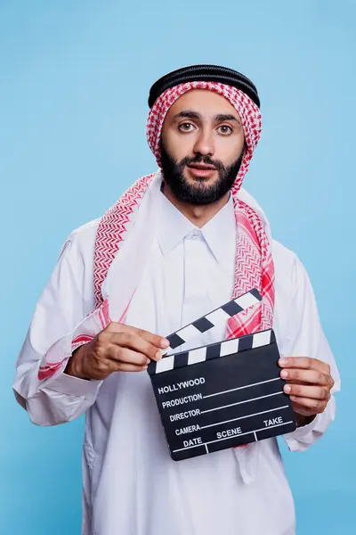 Movie scene director wearing traditional muslim clothes holding clapperboard and looking at camera. Arab dressed in islamic headscarf and thobe using clapboard studio portrait