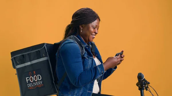 African american female exploring mobile phone while on bicycle ready for food delivery service. Young black woman embracing fast-paced economy by utilizing smartphone for navigation.