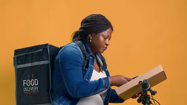 On her bicycle, african american courier delivers takeout orders while using wireless payment gadget for seamless transactions. Delivery woman pulls pos machine from pocket for contactless payment.