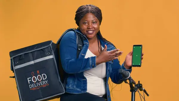 Lady with african american ethnicity showing cell phone with blank chromakey mockup template. Technology-driven delivery person grasps mobile phone with her left-hand displaying green screen.