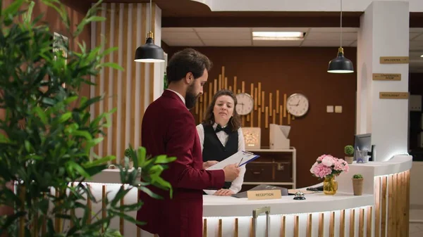 Gentleman signing registration papers at front desk, doing check in procedure for business trip accommodation at hotel. Receptionist woman welcoming businessman in lobby, friendly service.