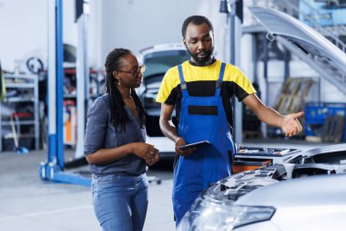 Araba servisindeki tamirci, tablet kullanan hasarlı araç için yeni kompresör kemeri istiyor. Çalışmayan otomobilin eski parçalarını değiştirmek için çevrimiçi bileşenleri arayan müşterinin yanındaki eğitimli teknisyen