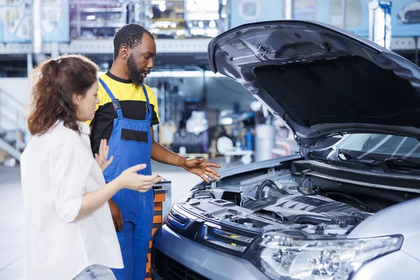 African american mechanic helping client with car maintenance in auto repair shop. Employee in garage facility looking over automobile parts with woman, mending her vehicle engine during inspection