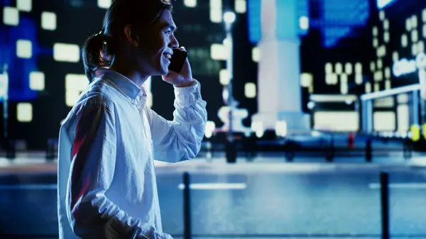 Asian man talking over the phone with wife using cellphone to feel less bored while strolling around dimly illuminated empty avenues at night, blurry cityscape background with bokeh lights