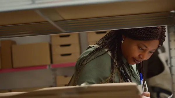 Close up shot of meticulous african american warehouse worker scanning cardboard boxes on shelves, updating merchandise labels info before sending parcels to be dispatched