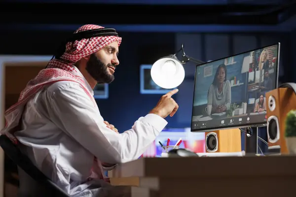 コンピュータでは ムスリムの男性がグループビデオ会話中に同僚の話を注意深く聞いています サイドビュー写真は 伝統的にオンラインミーティングを開催して服を着た中東の男性を示しています — ストック写真