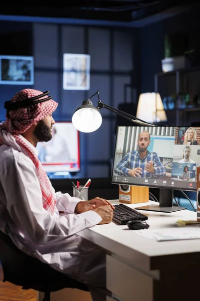 オフィスのテーブルでは 伝統的な服装を着たアラブ人がオンラインビデオミーティングに参加します イスラム教徒の男は コミュニケーションと研究のためにデスクトップPcを使用しながら同僚に細心の注意を払っています — ストック写真