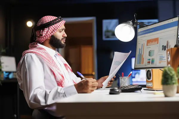 中東の男性フリーランサーは独立して働き アイデアを生み出し 現代のコンピュータ上でさまざまなオンラインタスクを管理しています 彼のPcモニター上の情報と彼のペーパーワークを比較するアラブの男 — ストック写真