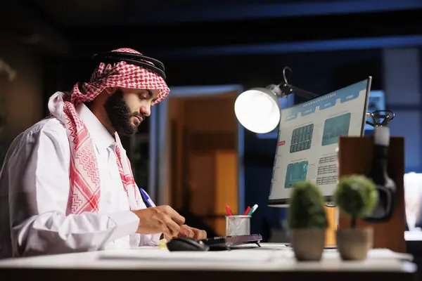 伝統的な服装のイスラム教徒のビジネスマンは 彼のコンピュータ画面からノートを下げるためにペンを使用します この画像はデスクトップPcとメモ帳で研究を行っている勤勉なアラブ人男性を描いています — ストック写真