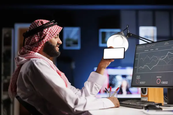 stock image An Arabic individual, wearing traditional clothing uses a mobile phone at an office desk. The isolated white screen offers space for customized content in an advertising or promotional setting.