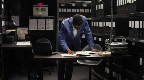 African american private investigator standing and cross-checking clues and evidence with laptop. Black policeman with criminology knowledge examines forensic files and classified research data.