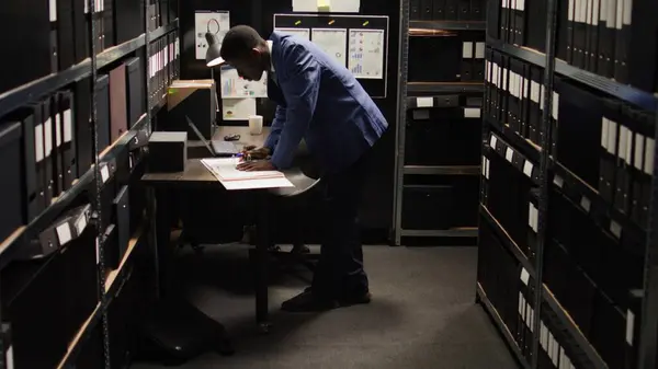 African American policeman carefully analyzes material, does records searches, and collects data from shelves and laptop. Male police officer standing, looking through case files on desk in office.