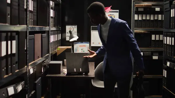 Male private investigator exits office room after packing his personal computer and closing it. African american police officer setting up his desk, organising paperwork, and carrying his laptop bag.