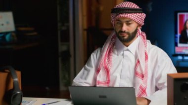 Evden çalışan, bilgisayarında yazı yazan, internette sörf yapan gülümseyen Arap serbest çalışanın portresi. Geleneksel giyinmiş Ortadoğulu adam dijital cihazda geziniyor, müşterilere e-posta gönderiyor
