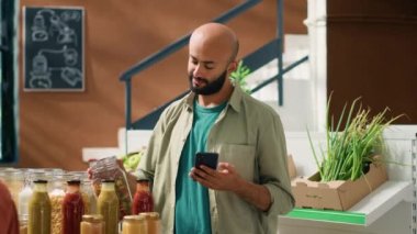 Ortadoğulu adam cam kaplarda saklanan ve akıllı telefondan organik ürünlerin faydalarını okuyan makarna listelerini kontrol ediyor. Vejetaryen müşteri, ekolojik bakkaliyeyi garantiliyor..
