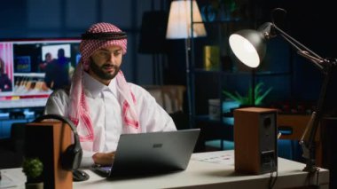 Mutlu Arap serbest çalışanın portresi evde çalışırken görevleri çözüyor, dizüstü bilgisayarda klavye kullanıyor. Ortadoğulu adam dijital cihaza bakıyor, ofis ortamındaki müşterilerin e-postalarını okuyor