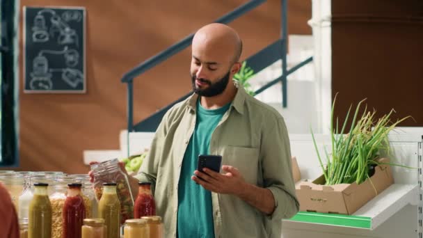 中東の男性は ガラス容器に保存されていないパスタの成分リストをチェックし スマートフォンで有機製品の利点を読みます エコ天然食料品についてのビーガンクライアントの確保 — ストック動画