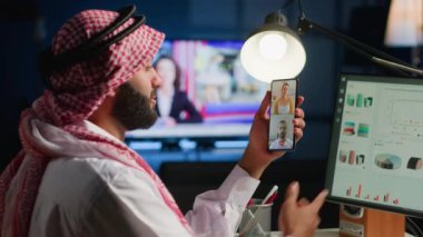 Müslüman tele-işçi ve çevrimiçi video çağrıları analitik istatistiksel veri setlerini loş aydınlatılmış dairede kontrol ediyorlar. Telekonferans toplantısında kilit performans göstergelerini optimize eden Arap çalışan