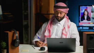 Ev ofisinin bilgisayarında çalışan gülümseyen Arap girişimcinin portresi, internette gezinen ve deftere yazan. Orta Doğulu adam telgraf çekerken kalem, not defteri ve dijital aygıt kullanıyor