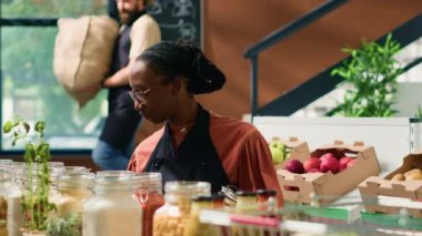 Raflarda taze hasat edilen organik ürünleri düzenleyen mutlu kadın bakkal. Gülümseyen küçük işletme sahibi yerel olarak yetiştirilen biyo-meyve, sebze, tahıl ve baharatları satıyor..