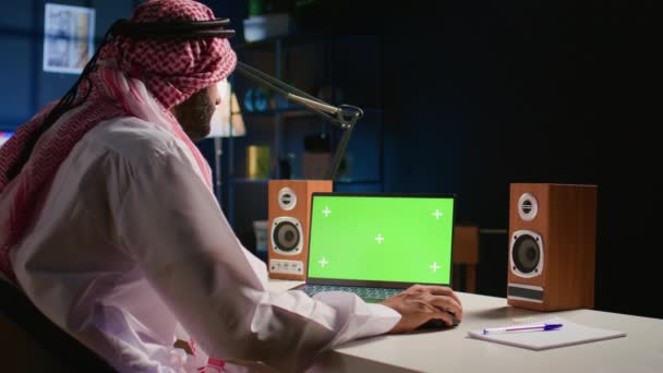 イスラム教徒の男性が緑色のスクリーンラップトップをスクロールし ソーシャルメディアフィードをチェックします バックグラウンドノイズとしてテレビ付きアパートオフィスのクロマキーデバイスからインターネットをサーフィンする自己雇用者 — ストック動画
