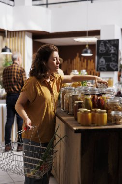 Elinde alışveriş sepeti olan bir kadın, sıfır atık mağazasından plastik kullanımı en aza indirmek için eko dostu ürünler alıyor. Besleyici yiyecekler arayan yeşil yaşayan bir müşteri.