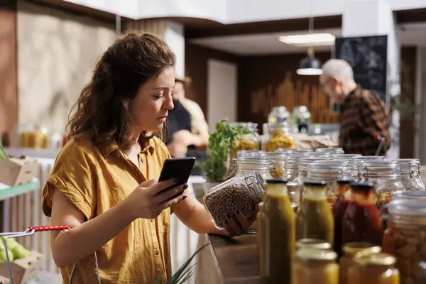 Woman in zero waste store using smartphone notes app to check shopping list. Meticulous customer looking to replenish pantry at home with organic chemicals free food essentials
